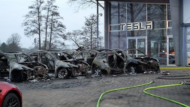 Several completely destroyed Teslas are in a row in front of a Tesla showroom entrance. Parts of the building are bubbled and roasted from the intensity of the EV fire.