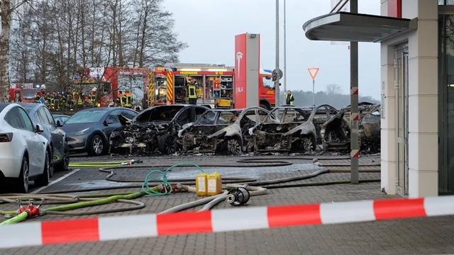 A fire crew is parked in the distance. A red Tesla sign looms over the burned out husks of Tesla sedans