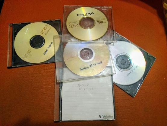 Cuatro CD-R y un DVD-R de datos, algunos en sus estuches plásticos, yacen sobre una superficie de color naranja oscuro.  Cada disco tiene escritura a mano que indica "Backup" y una fecha, entre el 2001 y 2008.