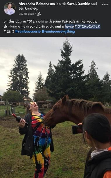 A woman in a colorful, tie-dye rainbow onesie is laughing hysterically as a brown horse affectionately nudges or "motorboats" her stomach. She's holding a bottle of wine in her left hand with the background revealing a grassy field with trees. Another person is visible taking pictures of the encounter