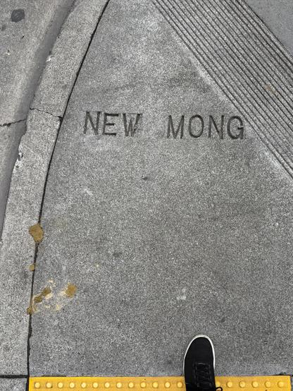Photograph of an urban #sidewalk street corner and yellow textured curb ramp. The camera’s perspective is looking straight down at the photographer’s foot and a street name stamped into the concrete. It reads:

"NEW MONG”