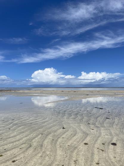 Uma cena de praia serena com padrões de areia que levam a águas calmas e rasas. O céu é azul vibrante com nuvens brancas espalhadas, refletindo a atmosfera tranquila.