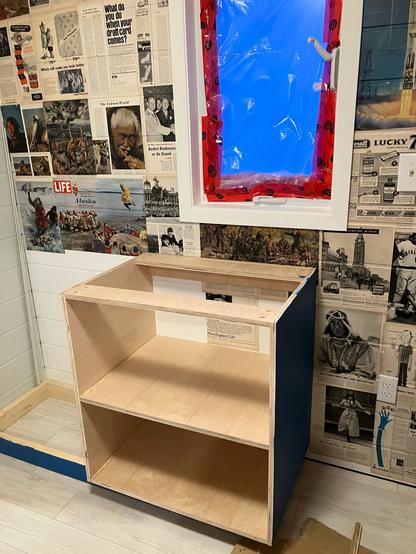 One half of a kitchen cabinet for the cabin sits on top of the kick.  The wall behind is plastered with pages from old life magazines.