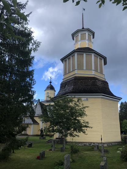 The photo shows the bell tower in the foreground with the church behind in a leafy cemetery setting. The church and tower are clapboard wooden buildings painted pale yellow. The bell tower is six-sided and has three tiers which get much smaller towards the top. Its window openings are boarded up. Photo copyright Coralie Mills 2024