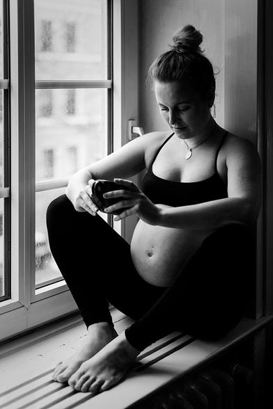 Eine schwangere Frau sitzt auf der Fensterbank, schaut nachdenklich zu ihrem Bauch und hält eine Tasse in der Hand.