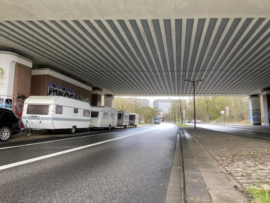 Vier heruntergekommene Wohnwagen stehen am Rand der Kieler Olshausenstraße unter der Bundesstraßenbrücke. Im Hintergrund sind die Gebäude der Universität zu sehen.