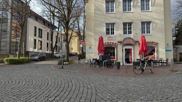 Das Bild zeigt einen kleinen Platz in der Osnabrücker Weststadt. Im Vordergrund befindet sich ein gepflasterter Bereich, der zu einem Café oder führt. Das Café hat rote Sonnenschirme und Tische im Freien. Im Hintergrund sind Gebäude zu sehen, eines davon größer und mit mehreren Fenstern. Die Szene wirkt ruhig und einladend.