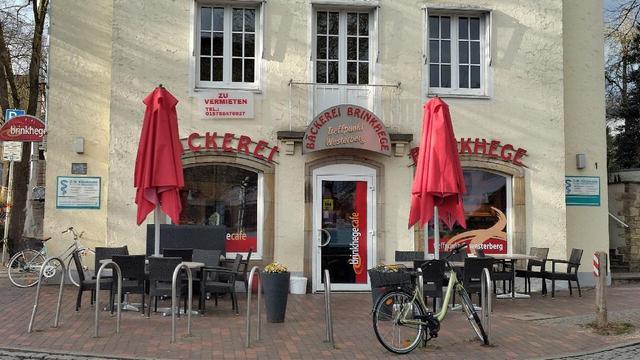 Das Bild zeigt einen kleinen Platz in der Osnabrücker Weststadt. Im Vordergrund befindet sich ein gepflasterter Bereich, der zu einem Café oder führt. Das Café hat rote Sonnenschirme und Tische im Freien. Im Hintergrund sind Gebäude zu sehen, eines davon größer und mit mehreren Fenstern. Über einem Fenster ist ein Schild angebracht: Zu vermieten. Die Szene wirkt ruhig und einladend.