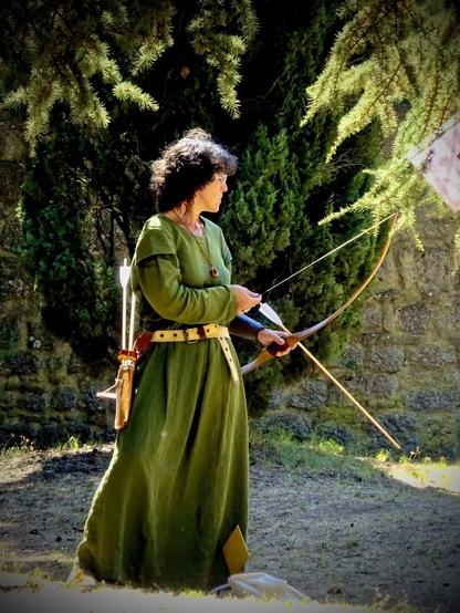 A woman dressed in historical attire, wearing a long green garment and a belt, is holding a bow in a natural setting. Behind her are trees and a stone wall. Several arrows are visible in a quiver attached to her belt.