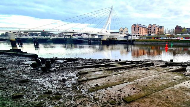The remains of a shipyard slipway on a foreshore with a modern bridge in the background.