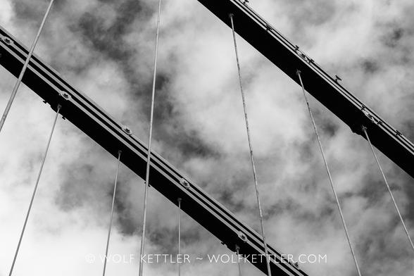 Black and white photograph looking up at two supporting cables of Clifton Suspension Bridge, running diagonally from top left to bottom right, parallel to each other. Thinner cables coming down vertically. Background is a dramatic, dark sky with white clouds.