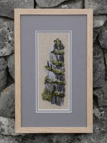 Stitched artwork depicting part of a slate quarry with bands of vegetation between the horizontal layers of striated rock. A tall image, framed with a grey mount to compliment the stone and a plain oak frame. Viewed face on against a stone wall