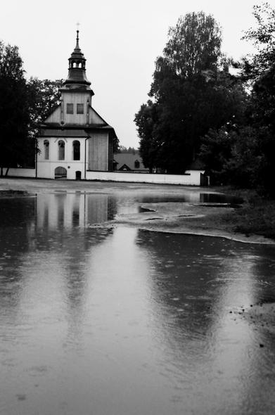 Dziedziniec drewnianego kościoła w ulewnym deszczu, na pierwszym planie okazała kałuża