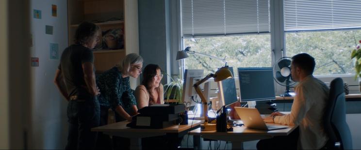 A group of scientists in a modern office is gathered around a computer looking at the screen.