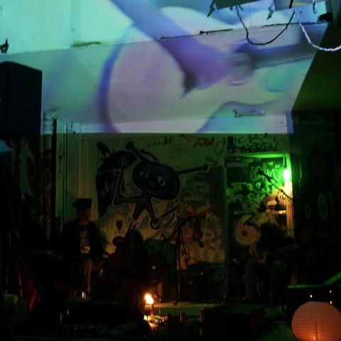 A psychedelic mélange - four dim figures beneath psychedelic light projections can be made out seated in front of a graffiti art wall, a projected figure on the roof playing a guitar. It's all a bit hard to comprehend! From left to right - Misbah, Daevid Allen, Kavi Samut, and myself playing guitar.