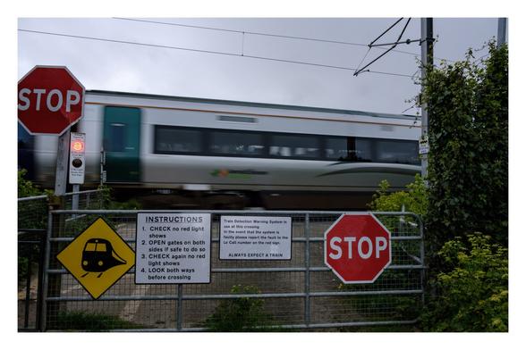 Foto im Querformat. Blick auf einen Bahnübergang. Es fährt gerade ein grauer Zug vorbei, durch die längere Belichtungszeit ist er leicht unscharf. Vor dem Bahnübergang ist ein geschlossenes Gitter, darauf sind mehrere stop Zeichen und Schilder. Darauf steht: 

INSTRUCTIONS
1. CHECK no red light shows
2. OPEN gates on both sides if safe to do so
3. CHECK again no red light shows
4. LOOK both ways before crossing
Train Detection Warning System in
use at this crossing.
In the event that the system is
faulty please report the fault to the
Lo Call number on the red sign.
ALWAYS EXPECT A TRAIN
STOP