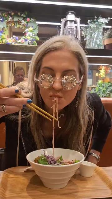Katherine enjoys a bowl of vegan ramen while wearing several pieces of Xover0 jewelry, including OPULENZ— the over-the-top eyeglasses created for your entertainment. Duane sits across from her and is visible in the mirror over her shoulder.