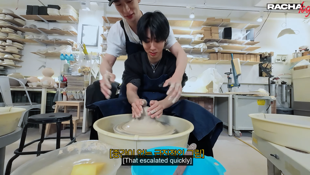 In a pottery studio, Han is attempting to do something with clay on a wheel. Hyunjin is behind him, leaning forward with his arms around him, to help guide him. Caption: [That escalated quickly]