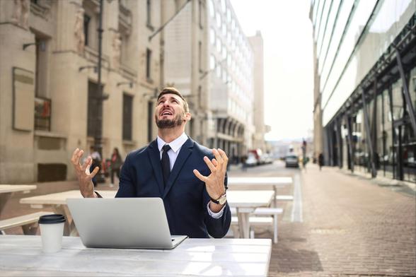 A man sitting at a table with his laptop in the middle of a city street.  He is looking up at the gray sky with a look of utter exasperation.