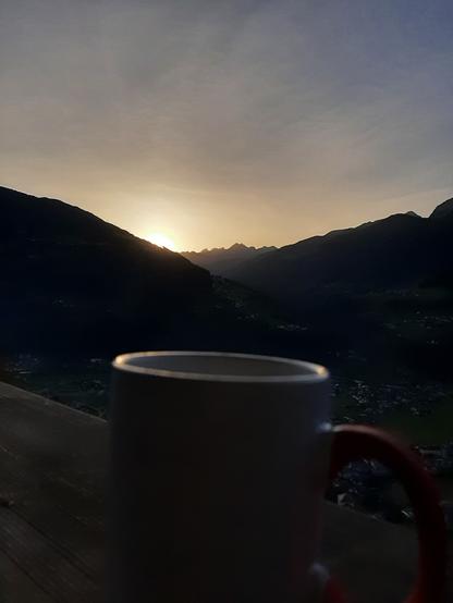 Das Bild zeigt das Zillertal bei Sonnenaufgang. Im Vordergrund befindet sich ein weißer Becher mit einem roten Griff, der auf einem Holztisch steht. Der Hintergrund ist von majestätischen Bergen geprägt, die in der Morgendämmerung dunkel und schattig erscheinen. 

Der Sonnenaufgang taucht den Himmel in warme, goldene und orangefarbene Töne, während der Sonnenball hinter den Bergen sichtbar ist. Im Tal unter den Bergen sind kleine Siedlungen oder Dörfer zu erkennen.

Text mit Unterstützung von @altbot