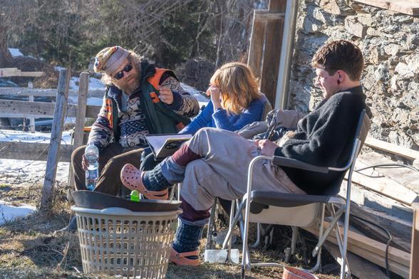 Wolfgang Lampl, Marlene Hauser und Simon Morzé sitzen in einer Drehpause vor eine Berghütte in der Sonne. Alle sind in dicke Winterklamotten gehüllt. Lampl der uns als einziger das Gesicht zuwendet trägt eine schwarze Sonnenbrille und eine Haube und in der linken Hand eine Plastikflasche Wasser.