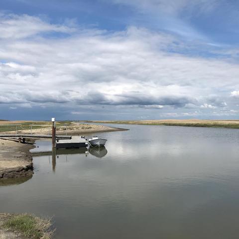 Das Bild zeigt eine ruhige Wasserlandschaft unter einem bewölkten Himmel. Ein kleiner Steg mit einem Boot liegt an einem sandigen Ufer. Das Wasser ist ruhig und reflektiert den Himmel, was eine friedliche und offene Szene erzeugt.