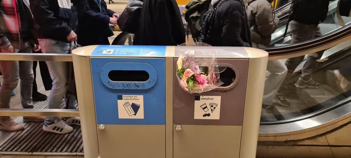A light grey trashcan with two seperate units next to an escalator full of partly visible people going up. The left unit has a blue front marked papers and magazine. The right unit has a dark grey front, is marked other trash and has a small, cheap looking boquet of pink flowers in clear foul sticking out of it.