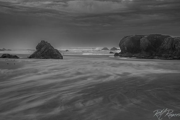 Black and white photograph of a wind swept beach on a stormy day. Large rocks rise from the shore, and the wind sweeps sand along the beach like smoke.