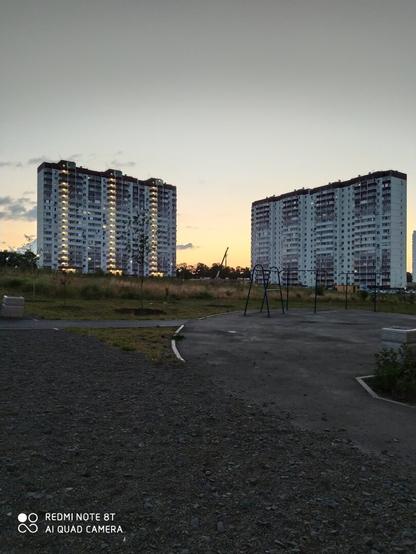 Два дома многоэтажных вдали, за ними жёлтое небо, с градиентным переходом от жёлтого к темно- серому. Переходные общие балконы между этажами светятся - там горят лампочки. А на переднем плане несколько дорожек посыпанных мелким щебнем и кусочек земли слева внизу