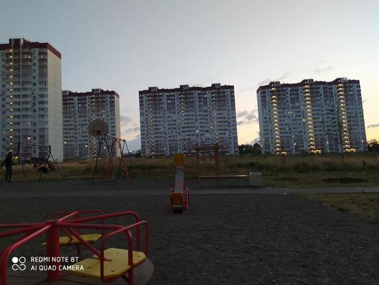 Несколько многоэтажек на фоне вечергего неба вдали. Небо ещё светлое. Вечер на переднем плане слева внизу видно кусочек карусели детской. А чуть дальше - качели.