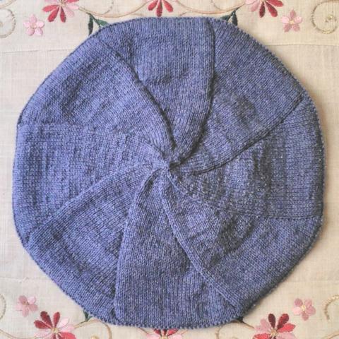Béret couleur bleu jean tricoté sur un fond écru dont on voit des broderies rouges et vertes en haut et en bas.