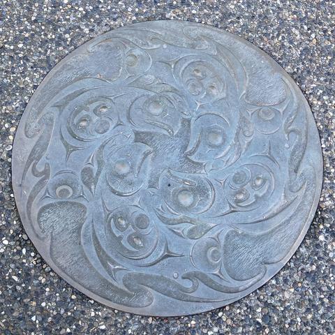 A silver manhole cover carved in Haida/Salish style, with four salmon making a ring, takes at the outside and faces curving in to the center, with a human-like face in the middle of each salmon's body.