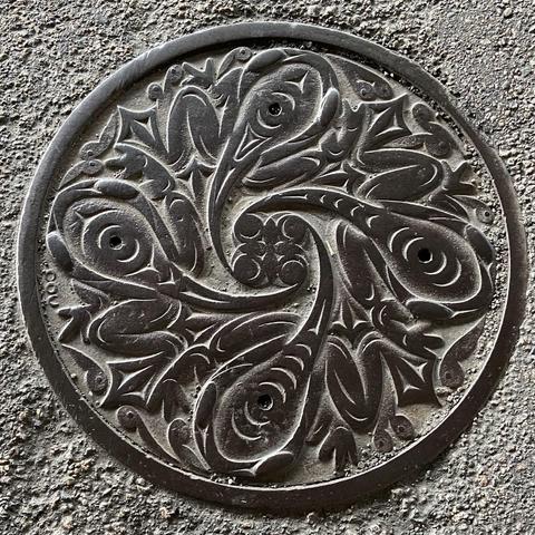 Black manhole cover in Haida/Salish style, with four frogs making a ring, faces to the outside of the circle