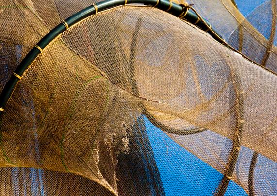 Nets on a large, historic sailing ship. There is a large black ring, you can see three quarters of it. The ring runs from the left, along the top of the photo to the bottom right. A brown net is attached to the ring all around. In some places you can look through it and see the blue sky. Behind it are more of these nets stretched. A net on the left is frayed. It looks like jute, but very thin. At the top right you can see a few more of those black rings with nets on them.