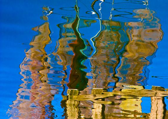 Reflection in the water of the 4 brown and dark red sails of the ship on a sky blue background. The photo is turned around, so you see them as in reality. The 4 mirrored sails are narrow and vertical. They are separate from each other and are distributed over the entire photo. At the bottom right of the photo the reflection of the yellow-brown slats of a wooden fence.