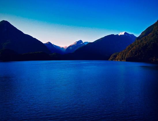 A serene fjord surrounded by dark, forested mountains with a clear blue sky and distant snow-capped peaks illuminated by the rising sun.