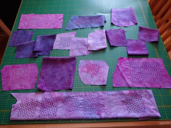 Seventeen small-to-medium square and rectangular pieces of cotton fabric dyed in shades of pink, purple, and blue, laid out on a cutting mat. Hitomezashi designs have been stitched on them in coordinating colours: crosses, squares, and fractal-looking designs, some regular and some random.
