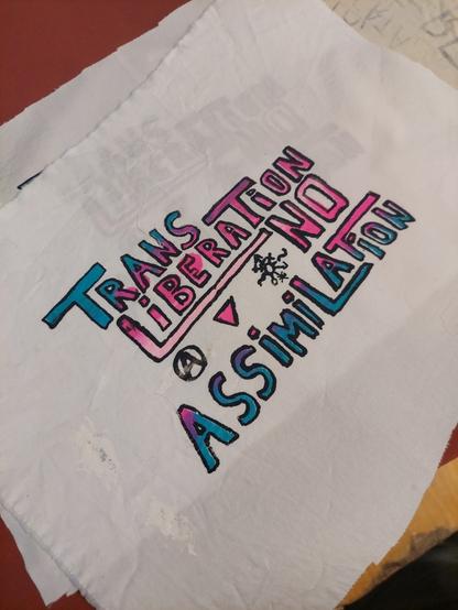 Trans Liberation patch, with letter filled-in in a trans flag gradient.