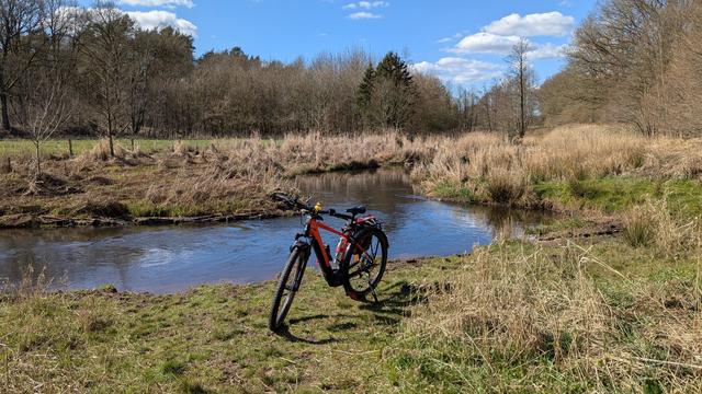 Blick in ein Autal, die Au mäandert durch die Landschaft. Im Hintergrund stehen einige Bäume. 
Im Vordergrund steht ein rotes Raleigh Dundee Mountainbike