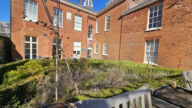 A photo of the rear of a Georgian redbrick building and garden containing a grade 2 listed garden which is overgrown. Touches of effort here and there but no consistency.
