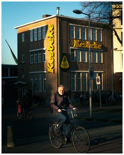 A person cycling in front of a building with a sign saying Kesbeke. The light lights the face of the cyclist and the sign.