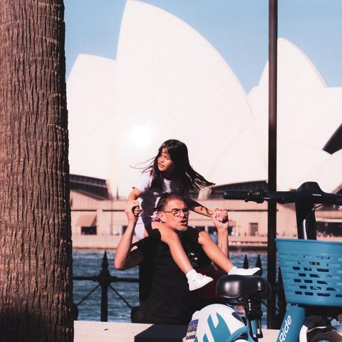 The image shows a man carrying a child on his shoulders near a waterfront. In the background is the Sydney Opera House. There is also a tree, an e-scooter, and a helmet nearby.