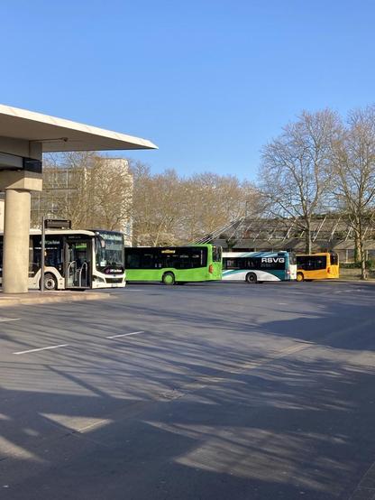 Sonne, blauer Himmel, links ragt das Dach des Busbahnhofs ins Bild, darunter 1 Bus in den Farben schwarz/weiss, dann reihen sich 3 weitere Busse an. Der nächste Bus ist grün, dann folgt ein weiß blaues Modell mit der Aufschrift Hybrid, zuletzt ist noch  ein Stück von einem orangefarbenen Bus zu sehen. Im Vordergrund Fahrbahnen in Asphaltgrau. Rechts in der Bildmitte stehen zwei unbelaubte Bäume.