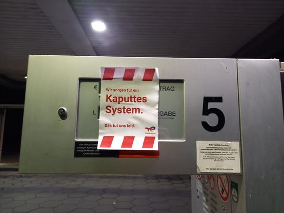Eine Zapfsäulenanzeige, die mit einem Poster in roter Schrift mit roten Streifen überklebt ist: "Kaputtest System." Darüber steht in klein: "Wir sorgen für ein", und darunter: "Das tut uns leid."