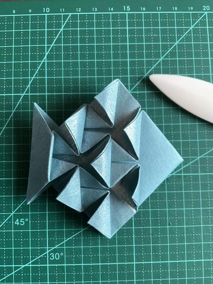 Poisson en origami, papier bleu métallisé, posé sur un tapis de coupe vert.