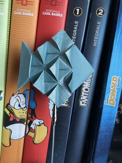 Poisson en origami, papier bleu métallisé, tenu par une pince devant une pile de BD Donald.