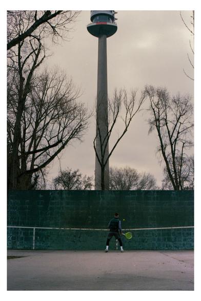 Color photograph of a scene in the Donaupark, Vienna. A person with a yellow tennis racket is wide-legged in anticipation of the yellow tennis ball, which is bouncing back from the practice wall. There are a few trees behind the wall and the Danube Tower can be seen a little further in the distance.