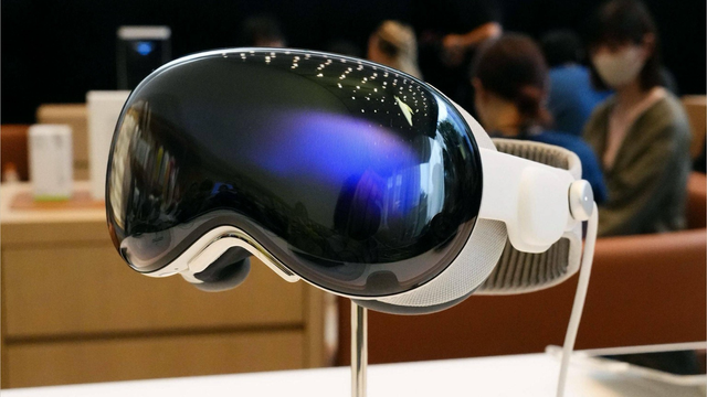 La imagen muestra un Apple Vision Pro sobre un soporte. El visor tiene una superficie reflectante oscura en la parte frontal. La parte posterior y la banda para la cabeza son blancas. El dispositivo está en exhibición sobre una mesa blanca. Al fondo, se ven personas y mobiliario, lo que sugiere que está en una tienda o en una sala de demostración.