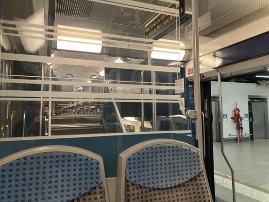 Interior view of a train. The colour scheme is grey and blue. It feels dim. We’re looking across the train, diagonally toward and open door. The platform is visible. We’d underground. In the middle distance we see the grey doors of two lavatorys.