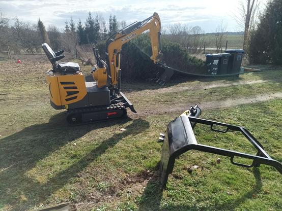 A yellow and grey tracked mini excavator from China. It's got a small dozer blade at the front and the arm has a digging bucket with teeth fitted. There's a seat with armrests and hydraulic joysticks, levers and pedals in front. The tracks are made from rubber and a red arrow points to where forward is (because the top can spin around and also face the other way). On the ground is a detached steel roll bar and sun roof and partially obscured by it, a wide sand bucket and a narrow trenching bucket.